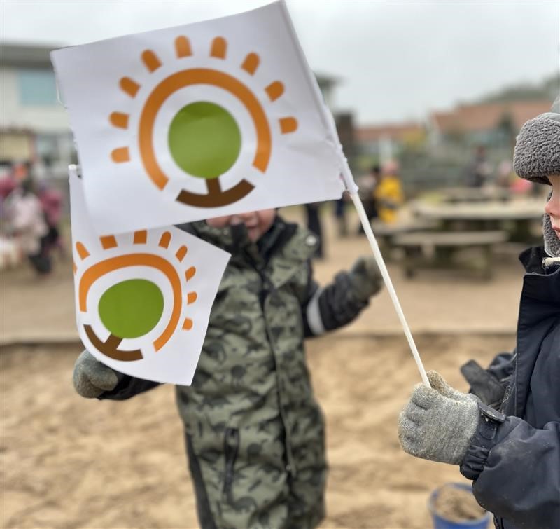 Ett barn utomhus vinkar med vita flaggor prydda med en orange och grön logotyp föreställande sol och träd. Barnen bär vinterkläder och vantar, med en förskolegård och picknickbord synliga i bakgrunden