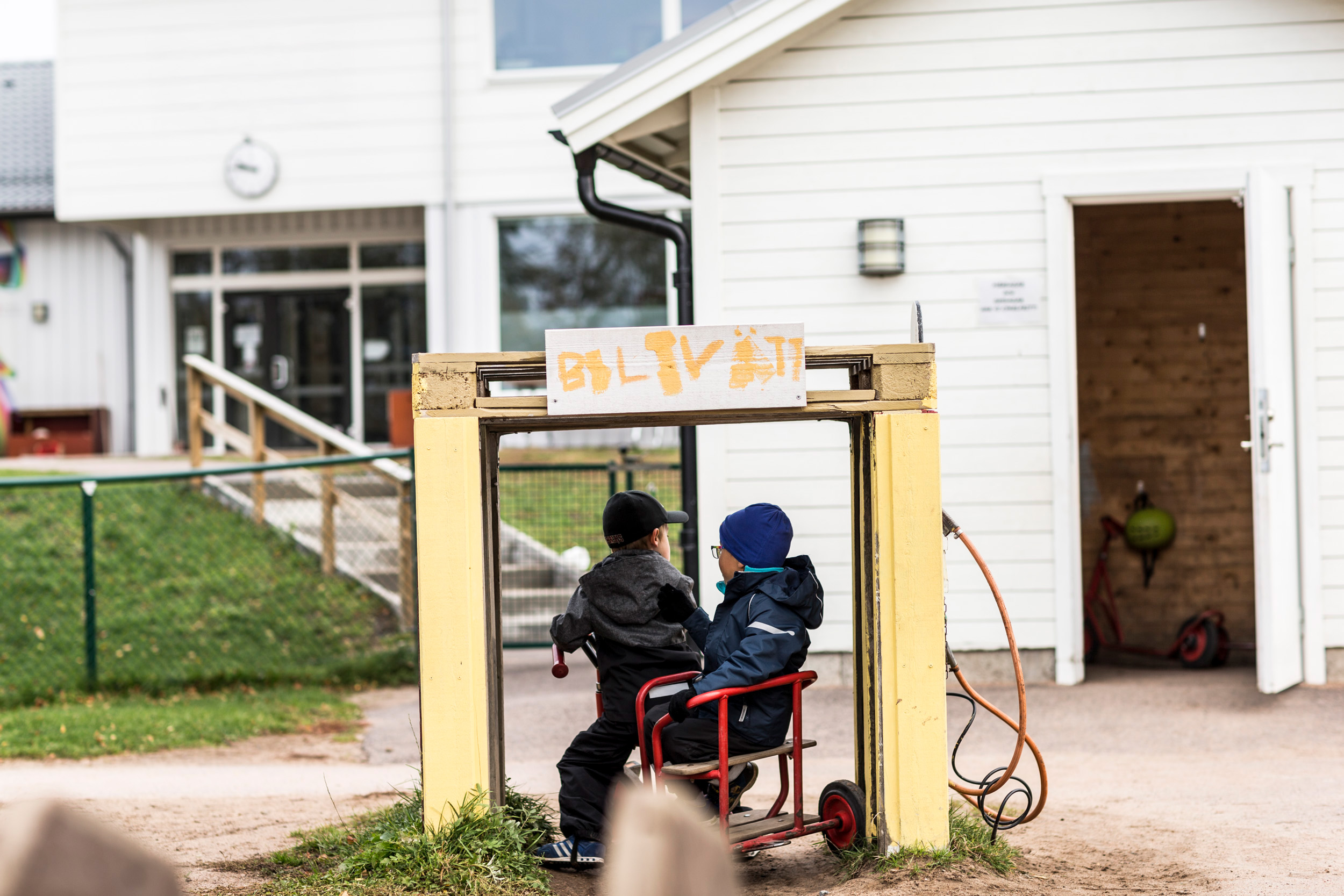 Två barn sitter på en röd trehjuling under en hemmagjord bilvättportal med skylten 'BILTVÄTT' målad i orange. I bakgrunden syns en vit förskolebyggnad med fönster och en ramp, omgiven av gräs och grus.