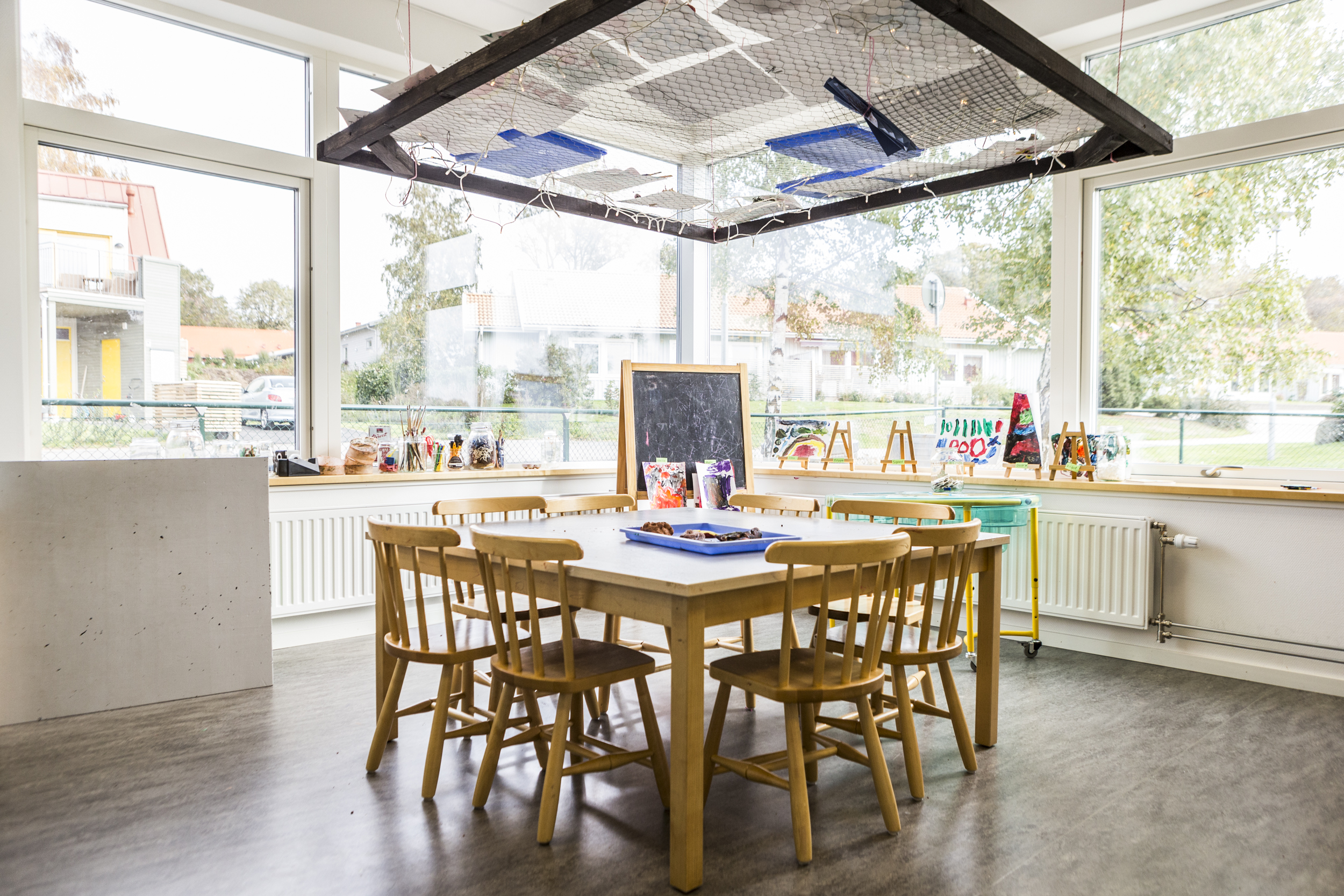 Ljust ateljérum på förskola med ett bord omgivet av trästolar, vid stora fönster med fönsterbänk full av pensel- och pysselmaterial. I bakgrunden syns små staffli med barnens målningar och en griffeltavla. I taket hänger en konstruktion av nät och lampor.