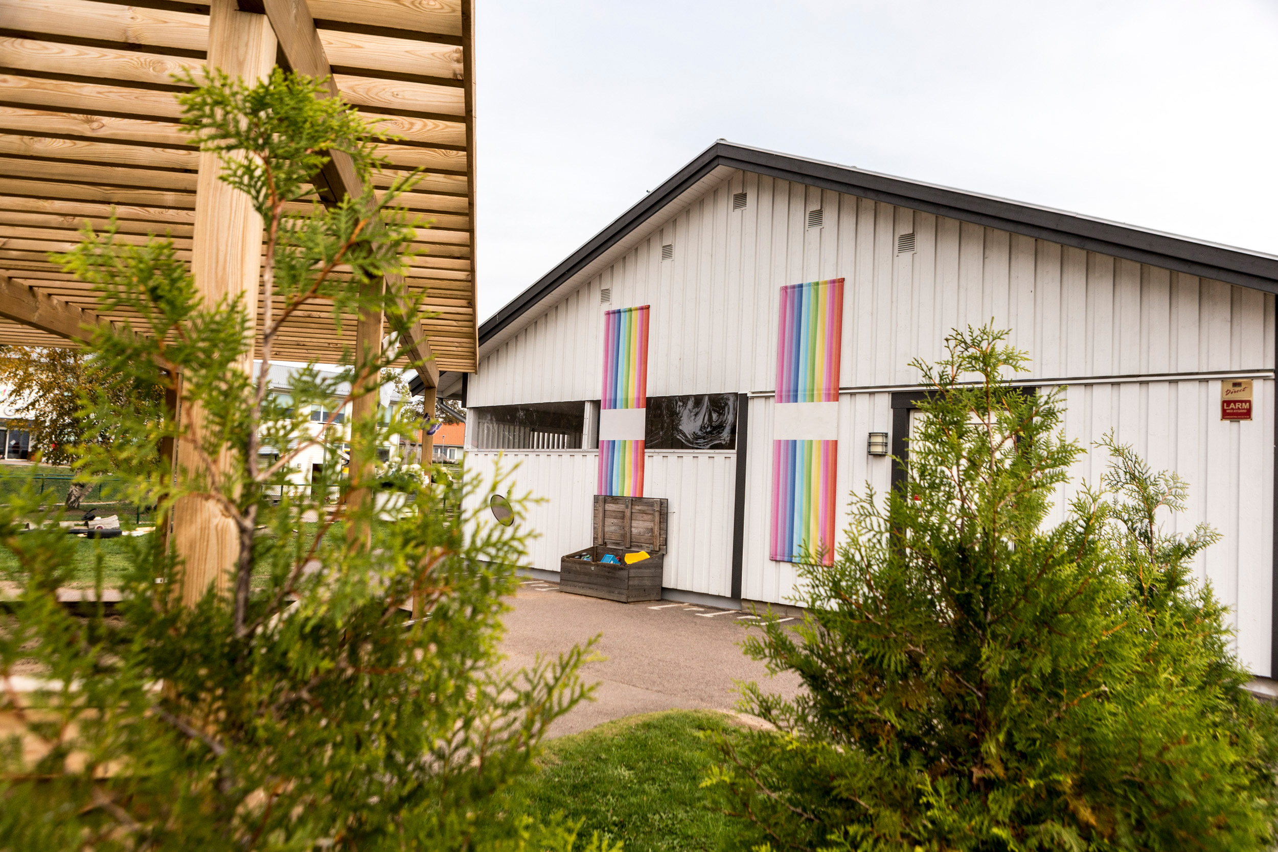 Vit förskolebyggnad med svart tak, dekorerad med två regnbågsfärgade banderoller på fasaden. Framför entrén står en trälåda med leksaker, och i förgrunden syns gröna buskar samt en trätäckt gångväg till vänster.