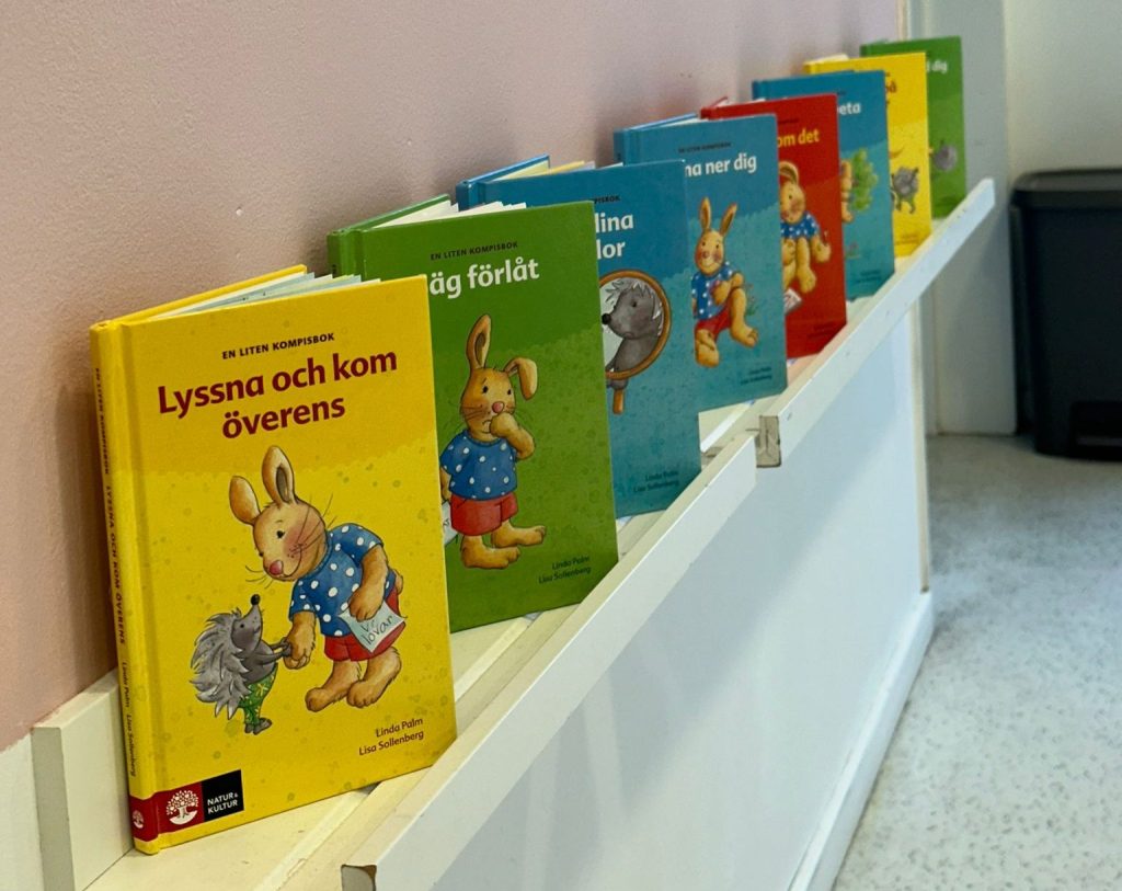 En rad böcker ur bokserien 'En liten kompisbok' med kanin- och igelkottsmotiv, uppställda på en vit vägghylla i en korridor. Böckerna har olika färgglada omslag i gult, grönt, blått och rött.
